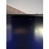 Borde superior de un monitor con pantalla oscura, dos pequeños puntos luminosos (amarillo, rojo), un reflejo claro a la derecha, superficie de madera y techo blanco al fondo