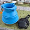 Zusammenklappbarer blauer Silikonbecher neben schwarzem Deckel auf grünem Tischset; im Hintergrund Metallbecher und Kunststoffdosen