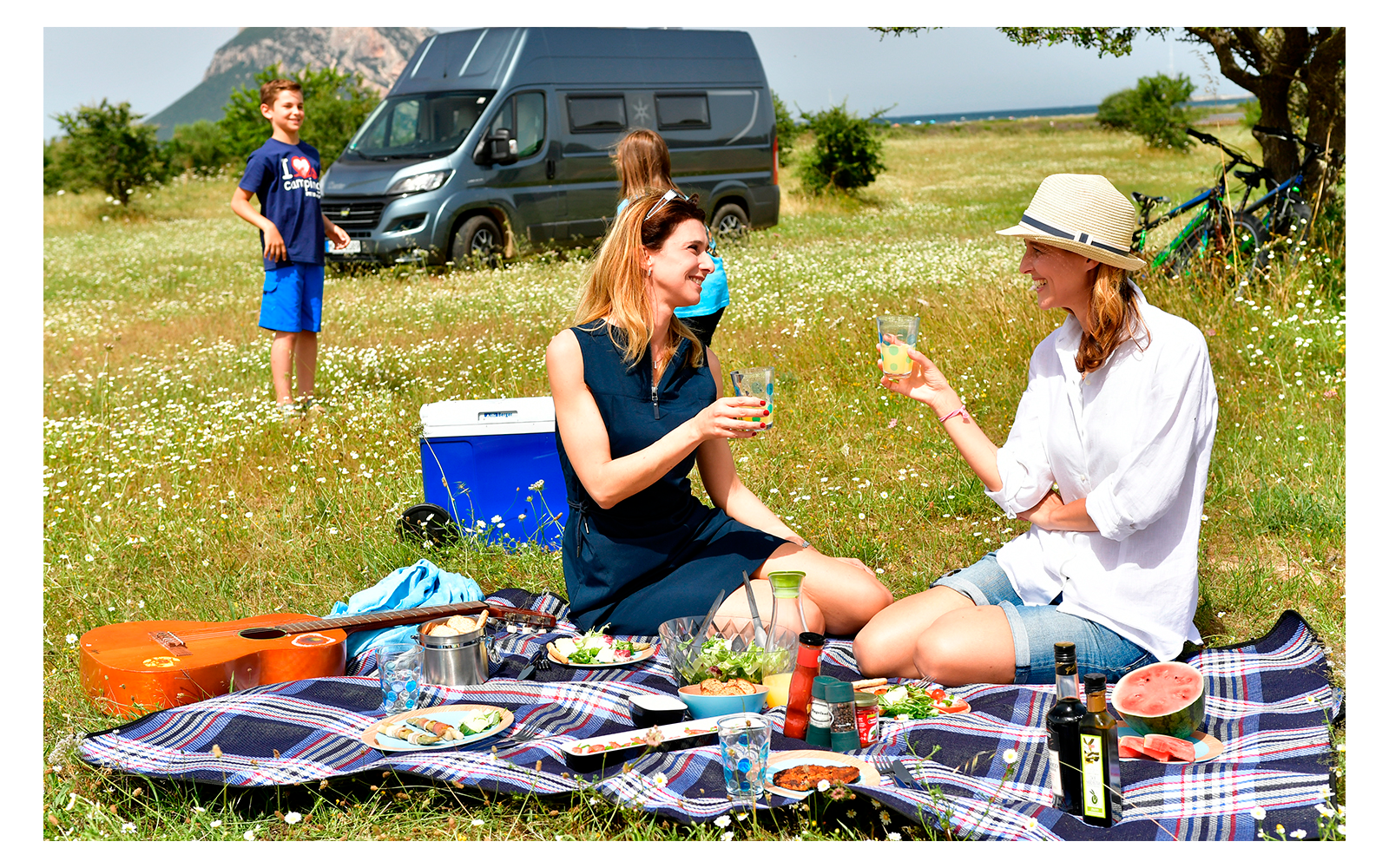 Drei Frauen beim Picknick auf einer Decke auf einer Wiese mit einem Camper im Hintergrund