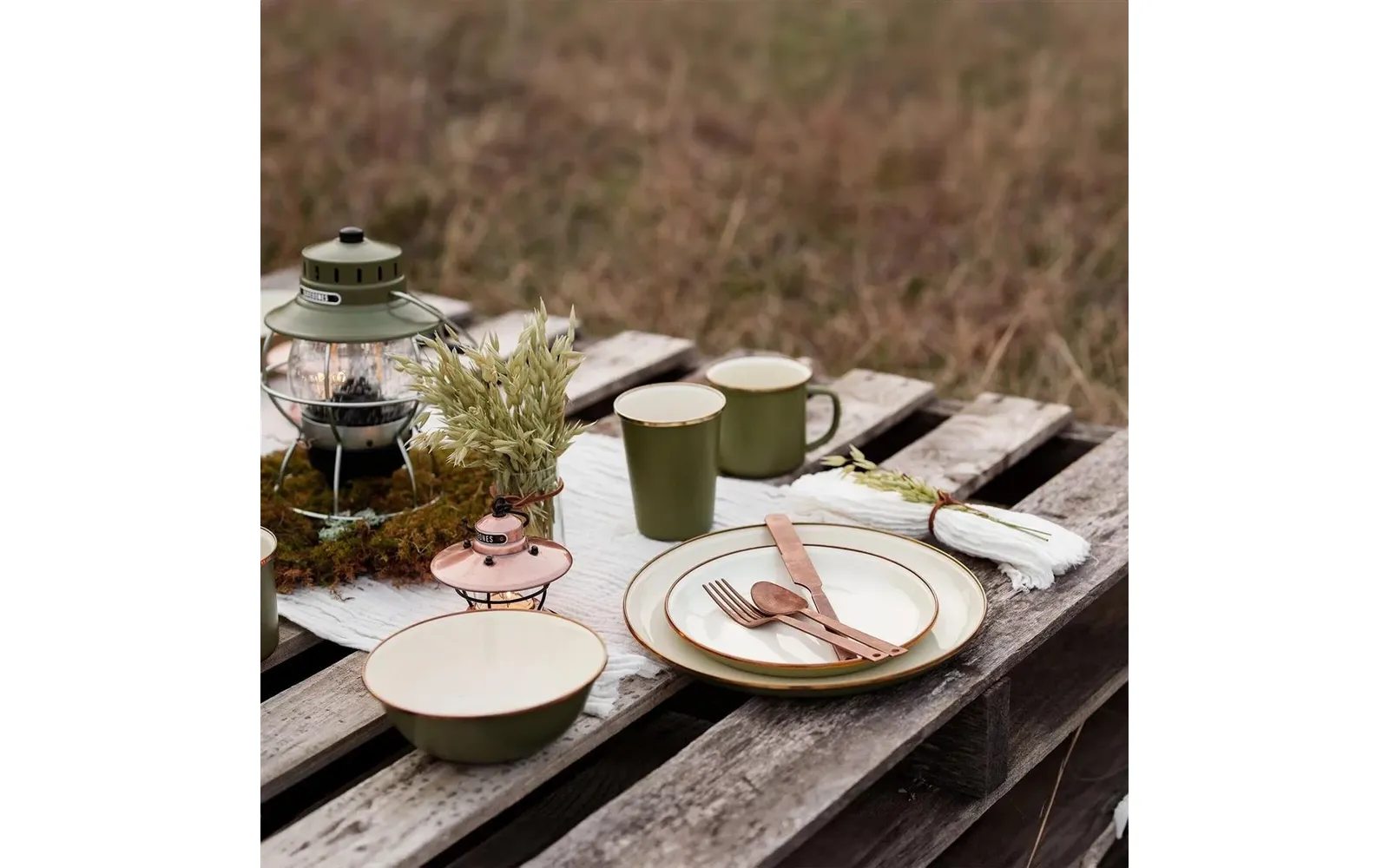 Houten pallet met twee keramische bekers, een keramisch bord met daarop een vork en een lepel, een kom, een lantaarn, een klein plantenpotje, een klein roze houdertje en gevouwen servetten; vage grasachtergrond