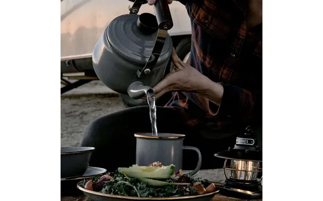 Hand gießt Wasser aus einem Teekessel in einen Metallbecher; im Vordergrund Teller mit Avocadohälfte und Salat, rechts eine Laterne