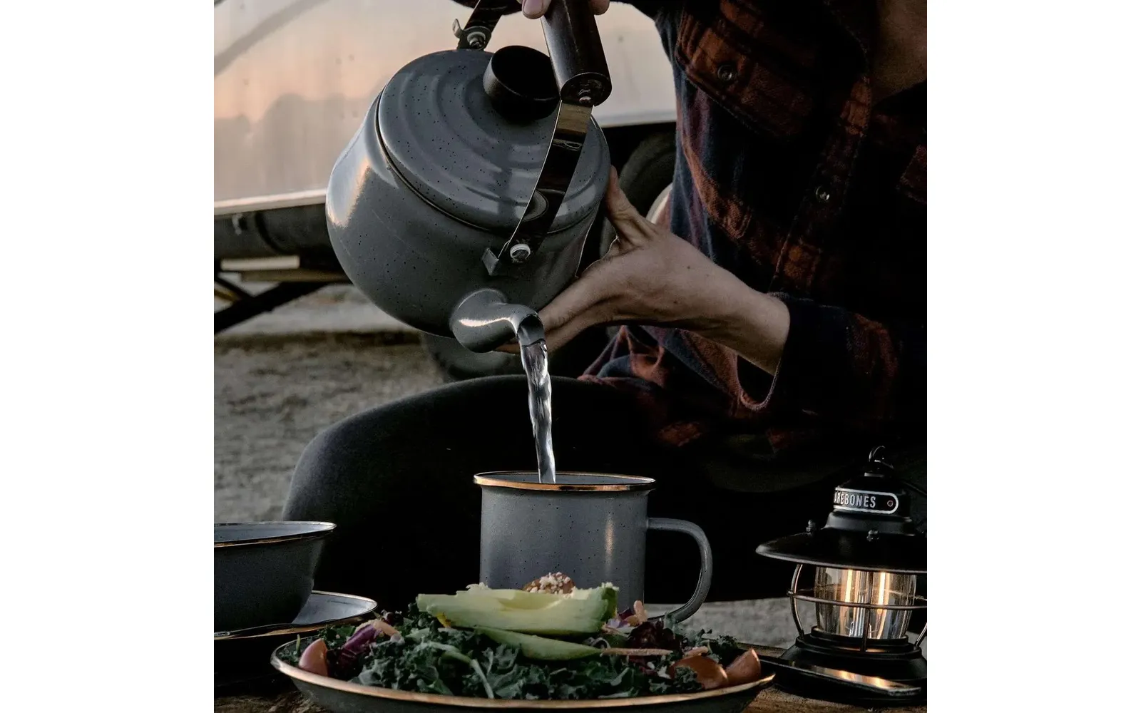 Hand gießt Wasser aus einem Teekessel in einen Metallbecher; im Vordergrund Teller mit Avocadohälfte und Salat, rechts eine Laterne