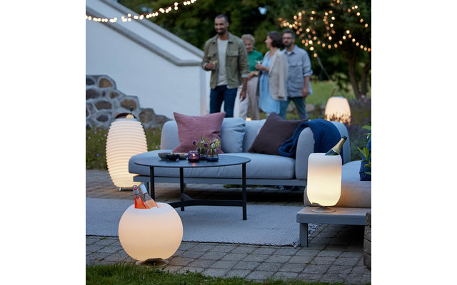 Außenbereich mit Sofa und Kissen, rundem Tisch, mehreren leuchtenden Lampen, Lichterkette und vier Personen im Hintergrund
