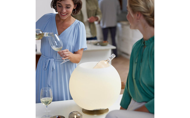 Zwei Frauen an einem Tisch; eine gießt Weißwein in ein Weinglas, die andere sitzt daneben. Auf dem Tisch steht eine beleuchtete Kugel mit einer darin liegenden Weinflasche, zwei Gläser und kleine Schalen