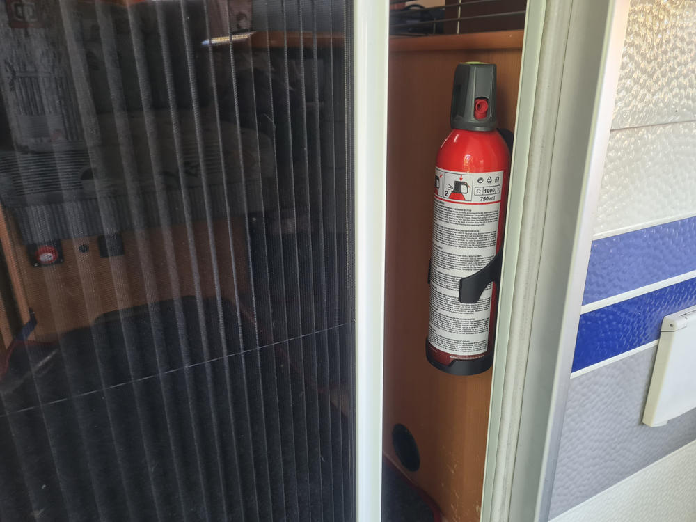 Roter Feuerlöscher mit Etikett und schwarzem Halteband an einer hellbraunen Holzplatte; links ein dunkler, gefalteter Vorhang; rechts ein weißer Türrahmen und eine weiße Wand mit blauem Streifen und kleinem Schalter