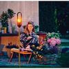 Person sitzt auf einem Stuhl im Garten, umgeben von Blumen, Pflanzen, einem Korb und einer stehenden Lampe