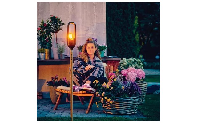 Person sitzt auf einem Stuhl im Garten, umgeben von Blumen, Pflanzen, einem Korb und einer stehenden Lampe