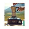 Two hands lift the lid of a stovetop espresso maker on a portable gas burner; blurred landscape in the background