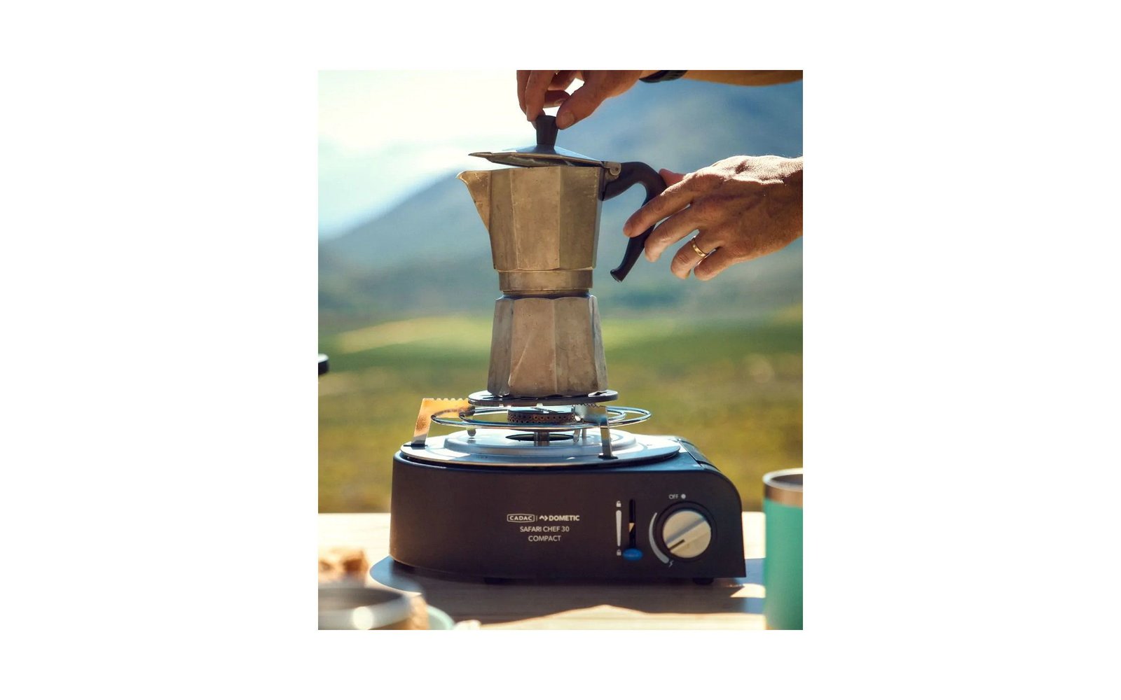 Two hands lift the lid of a stovetop espresso maker on a portable gas burner; blurred landscape in the background