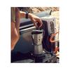 Hand places a moka pot on a portable camping gas stove in the open tailgate area of a vehicle; sandy rocks in the background
