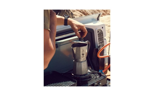 Hand places a moka pot on a portable camping gas stove in the open tailgate area of a vehicle; sandy rocks in the background
